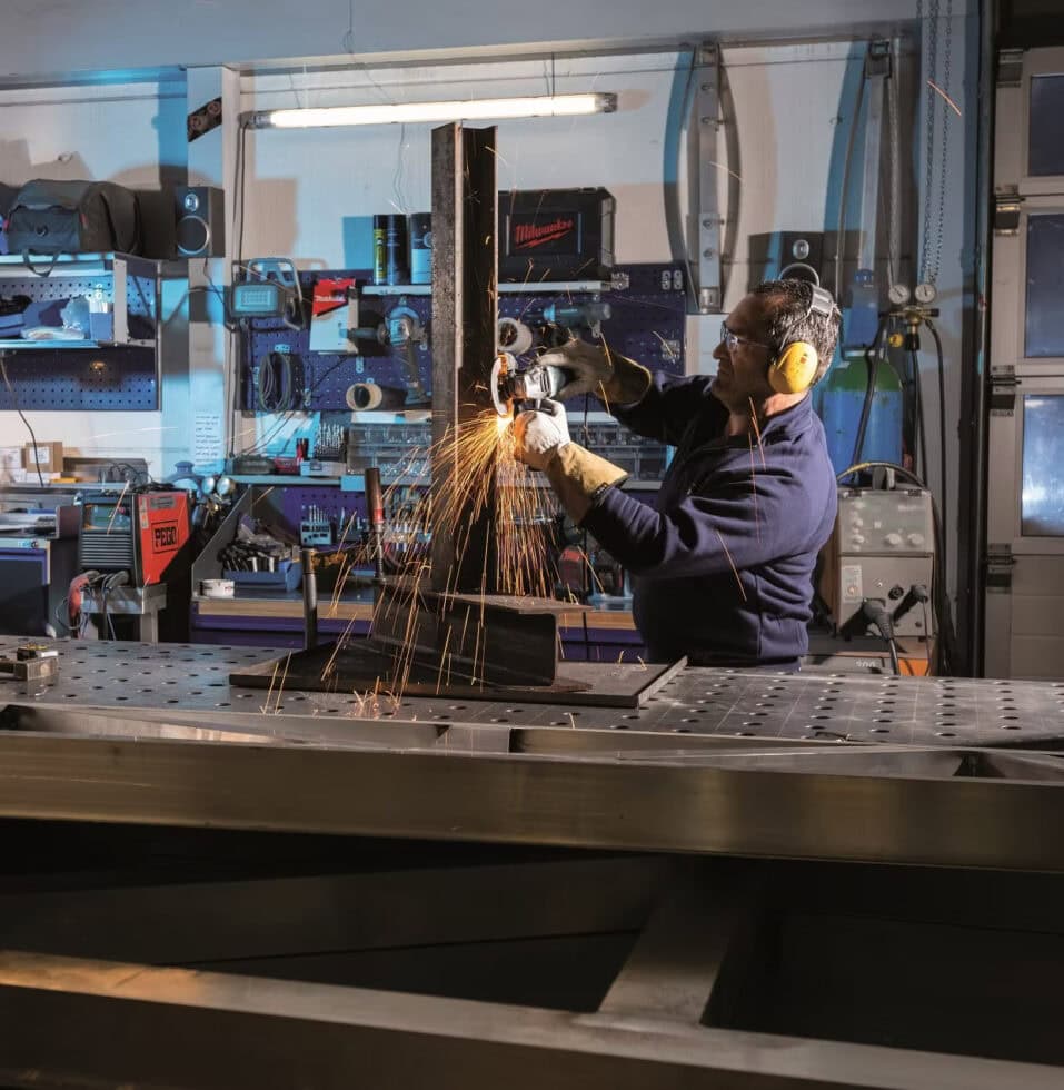 0edaf8_663168dba25c492b9bec451c839346f5~mv2 (1) Eine Person mit Gehörschutz und Handschuhen arbeitet in einer Werkstatt mit einem Winkelschleifer an einem Metallbalken, wobei beim Schleifen helle Funken fliegen.