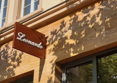 Ein braunes Schild mit dem Wort "Leonardo" in weißer Schrift ist an einem hellbraunen Steingebäude angebracht, das von Bäumen beschattet wird.