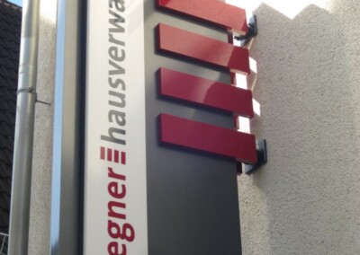 Vertikales Schild an einer Hauswand mit der Aufschrift "wegner hausverwaltung" mit roten und grauen Designelementen; Branding von ASSION Lichtwerbung.