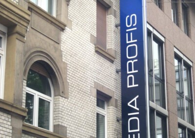 Auf dem vertikalen Schild an der Fassade steht "MEDIA PROFIS", darüber ein "mp"-Logo; das Gebäude weist architektonische Elemente aus Ziegeln und Stein auf.