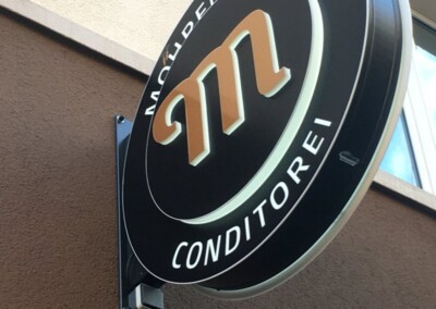 Auf einem runden Schild, das an einer Gebäudefassade angebracht ist, steht "Mohrenköpfle Conditorei" mit einem stilisierten orangefarbenen "m" und der Marke "ASSION Lichtwerbung" in der Ecke.