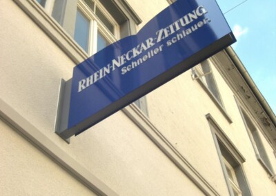 Blaues Schild für "RHEIN-NECKAR-ZEITUNG Schneller schlauer.", montiert an der Außenseite eines hellen Gebäudes mit Fenstern.