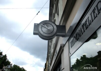 Ein schwarzes und silbernes Uhrenschild mit der Nummer 19 ist an der Außenwand über einem Schaufenster in einer Stadtstraße angebracht.
