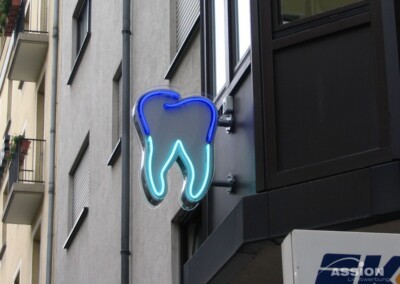 Eine Leuchtreklame in Form eines Zahns ist an der Außenwand eines Gebäudes angebracht und weist auf eine Zahnarztpraxis hin.