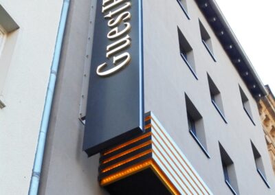 Eine Gebäudefassade mit einem vertikalen Schild, auf dem in großen Buchstaben "GuestHouse" steht, neben mehreren rechteckigen Fenstern.