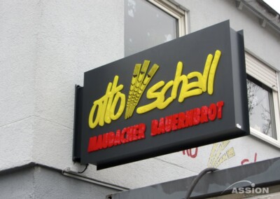 Ein Schild an einem Gebäude trägt die Aufschrift "otto schell MAIDACHER BAUERNBROT" mit einer Weizengrafik und erhabenen gelben und roten Buchstaben.