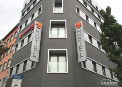 Ein modernes graues Hotelgebäude mit mehreren Fenstern und zwei vertikalen Schildern mit der Aufschrift "HOTEL" an der Fassade. Im Vordergrund sind ein Parkplatzschild und Bäume zu sehen.