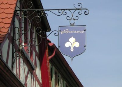 Ein dekoratives schmiedeeisernes Schild mit einer Lilienblüte und dem Wort "Renaissance" hängt vor einem traditionellen Fachwerkhaus.