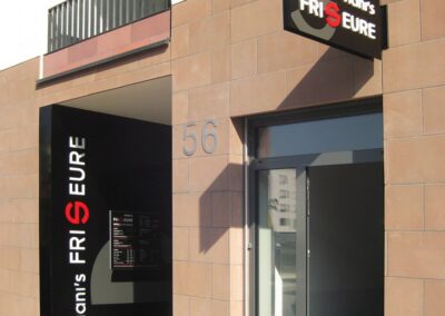 Ein moderner Friseursalon mit schwarzer Beschilderung und der Aufschrift "viani's FRISEURE" in weißen und roten Buchstaben über der Tür und an der Seitenwand. Die Hausnummer 56 ist sichtbar.