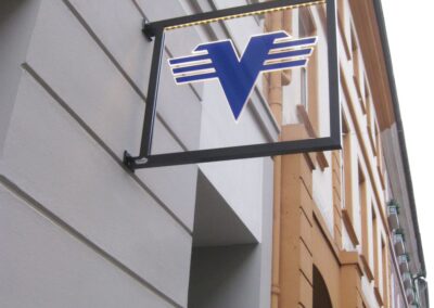 Ein quadratisches Schild mit einem blauen V und horizontalen Streifen ist an der Außenwand eines Gebäudes an einer Stadtstraße angebracht.