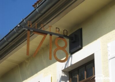 Metallschild mit der Aufschrift "BISTRO 1718", das an der Außenwand eines hellgelben Gebäudes über einer Tür angebracht ist.