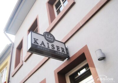 Ein rechteckiges Schild mit der Aufschrift "Kaiser Hotel Shop" ist an der Außenwand eines Gebäudes mit beigen Wänden und roter Umrandung der Fenster angebracht.