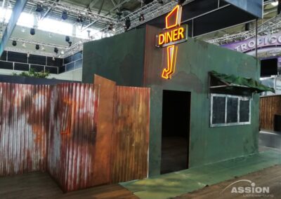 Ein kleines, verwittertes Gebäude mit einem Neonschild "Diner" und Wellblechwänden steht im Inneren eines großen Veranstaltungsraums.