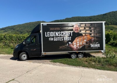 Ein Bäckerei-Lieferwagen, der an einem Weinberg geparkt ist, zeigt eine Werbung mit einem lächelnden Bäcker, der ein Brot in der Hand hält, und einem deutschen Text, der für Qualitätsbrot wirbt.