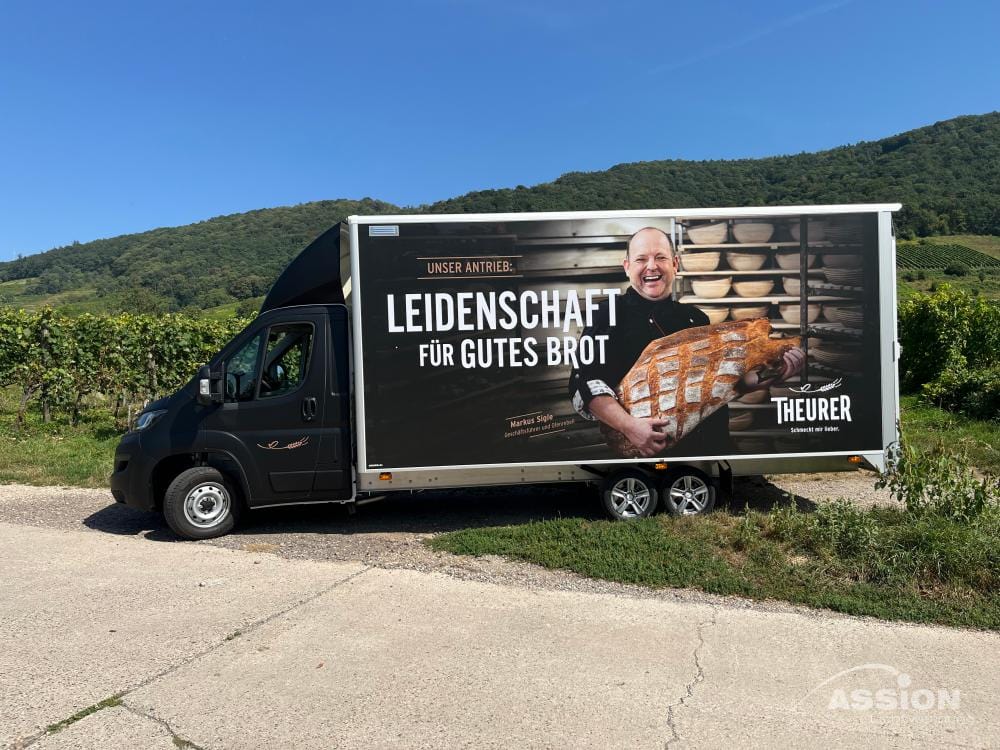 Autobeschriftung (1) XXX Ein Bäckerei-Lieferwagen, der an einem Weinberg geparkt ist, zeigt eine Werbung mit einem lächelnden Bäcker, der ein Brot in der Hand hält, und einem deutschen Text, der für Qualitätsbrot wirbt.