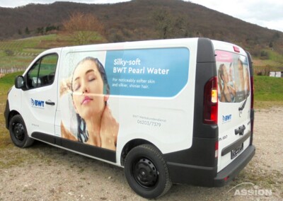 Ein weißer Lieferwagen mit BWT-Schriftzug und einer Werbeanzeige, die eine duschende Frau zeigt und für "Seidenweiches BWT-Perlwasser" für weichere Haut und glänzenderes Haar wirbt, parkt im Freien.