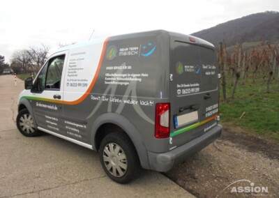 Ein grauer Lieferwagen mit dem Schriftzug "REHA TEAM MAISCH", der auf einer Landstraße neben Wiesen und Hügeln geparkt ist und auf dem an den Seiten und am Heck Kontaktinformationen und Dienstleistungen angegeben sind.