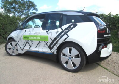 Ein weißer BMW i3 mit dem Schriftzug der Kraus Immobilien GmbH und geometrischen Grafiken ist auf einer Schotterfläche neben einer Grünfläche geparkt.