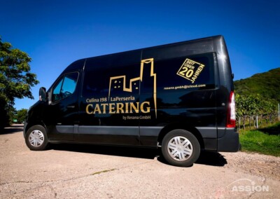 Ein schwarzer Lieferwagen mit goldenem Firmenlogo und Kontaktdaten steht auf einer Landstraße in der Nähe von Grünflächen und blauem Himmel.
