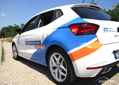 Ein weißer Schrägheckwagen mit blauer und oranger Aufschrift und dem Schriftzug der VR Bank Rhein-Neckar eG parkt auf einem Schotterweg neben einer Grünfläche.