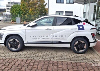 Ein weißer Geländewagen mit dem Schriftzug "KANZLEI GEORGIADIS Wirtschaftsprüfung - Steuerberatung" parkt vor einem Gebäude mit Glasfenstern.