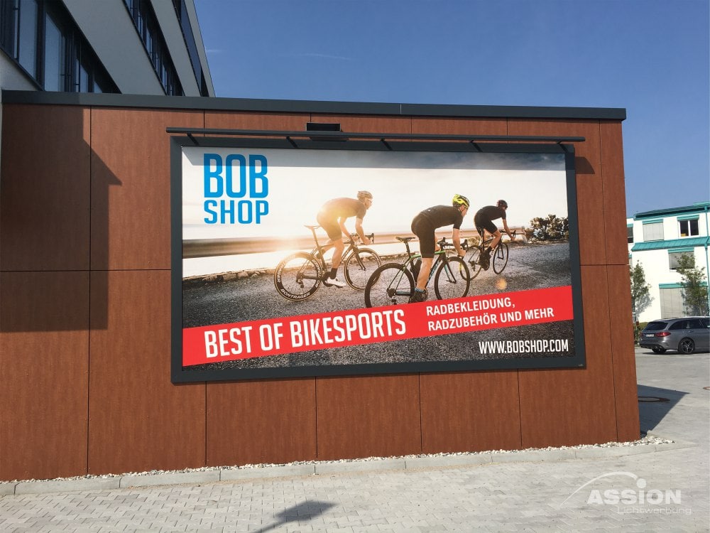 Banner — Fahnen (1) XXX Großes Außenbanner auf einem Gebäude zeigt Radfahrer auf einer Straße, die für "BOB SHOP" und "Best of Bikesports" werben, mit Website und deutschem Text über Fahrradbekleidung, Zubehör und sportliche Fahnen.
