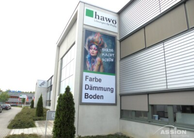 Ein modernes Gebäude mit einem großen vertikalen Schild mit einem bunten Porträt, deutschem Text und dem Firmennamen "hawo Lösungen für Profis" darüber.