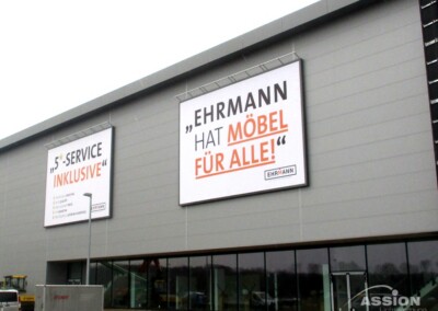 Große Schilder an der Außenseite eines modernen grauen Gebäudes werben für Möbel und Dienstleistungen von Ehrmann, mit fetter deutscher Schrift und orangefarbenen Highlights.