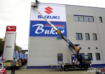 Ein Arbeiter auf einer erhöhten Plattform bringt ein großes Suzuki-Schild an der Außenwand eines Autohauses an.