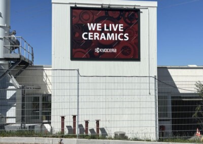 Auf einem großen Schild an einem weißen Industriegebäude steht "We Live Ceramics Kyocera". Davor steht ein Metallzaun und darüber ein strahlend blauer Himmel.