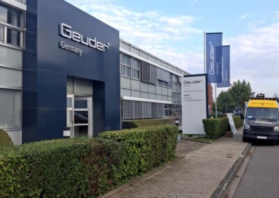Außenansicht des Bürogebäudes von Geuder Germany mit Firmenschildern, Hecken, einem Gehweg und einem geparkten Lieferwagen auf der Straße.