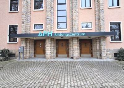 Außenansicht des Gebäudes des APH Bildungszentrums mit drei Holztüren, Steinsäulen und einem Schild über dem Eingang.