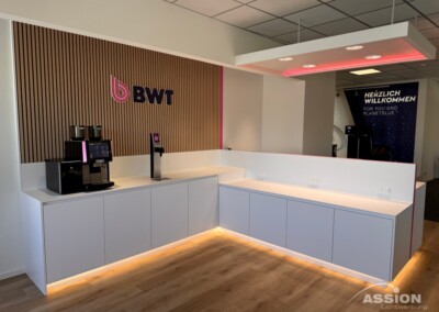 Moderne Büro-Kaffeestation mit Kaffeemaschinen, weißen Schränken, Holzfußboden und BWT-Logo auf einer Lattenwand. Deckenleuchten und Unterschrankbeleuchtung erhellen den Bereich.