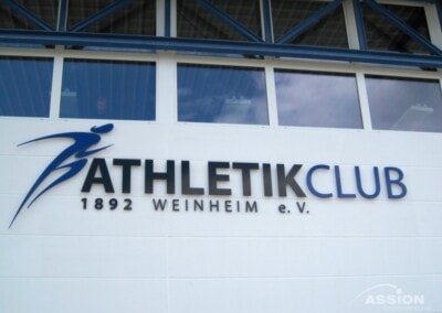 Außenwand mit dem Schild "ATHLETIKCLUB 1892 WEINHEIM e.V." und einem blauen abstrakten Logo neben dem Text. Über dem Schild sind die Fenster und Stahlträger sichtbar.