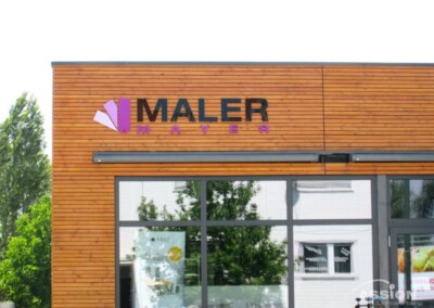 Eine hölzerne Gebäudefassade mit großen Fenstern zeigt ein lila-schwarzes "Maler Mayer"-Schild und ein lila fächerförmiges Logo.