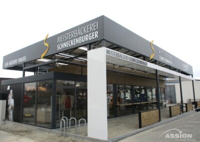 Modernes Bäckereigebäude mit Glaswänden und der Aufschrift "Meisterbäckerei Schneckenburger"; leerer Außenbereich sichtbar.