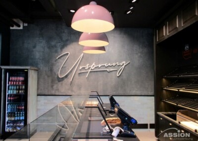 Modernes Bäckereiinterieur mit leeren Vitrinen, drei Hängelampen, einem Getränkekühlschrank und dem Schriftzug "Ursprung" an der Betonwand.