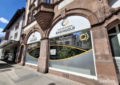 Straßenansicht des Rheingold-Büros mit großen Glasfenstern, auf denen das Firmenlogo und ein Text über den 24-Stunden-Hilfsdienst zu sehen sind.
