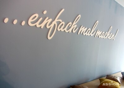 Auf einer grauen Wand steht in weißer Schrift "...einfach mal machen!" über einer Bank mit Kissen; das Logo "ASSION Lichtwerbung" befindet sich in der unteren rechten Ecke.