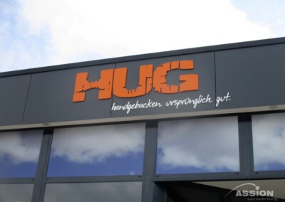 Eine Gebäudefassade mit einem orangefarbenen "HUG"-Schild und der Aufschrift "handgebacken. ursprünglich. gut." in weißer Schrift darunter. Der bewölkte Himmel spiegelt sich in den Fenstern.