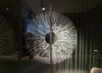 Großes, rundes Wandbild mit verzweigtem, weißem Muster, das an eine Iris erinnert, montiert an einer Glaswand in einem modernen Raum. Das Logo "ASSION Lichtwerbung" ist in der Ecke sichtbar.