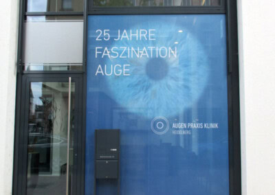 Großes blaues Schaufenster mit einer Augengrafik und deutschem Text: "25 Jahre Faszination Auge" in der Augen Praxis Klinik, Heidelberg. Vor dem Schaufenster befindet sich ein Briefkasten.