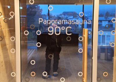 Glastür mit der Aufschrift "Panoramasauna 90°C" mit kreisförmigen Motiven; eine Person spiegelt sich im Glas; Innenraum aus Holz und Außenansicht im Hintergrund.
