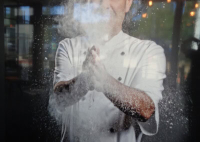 Ein Koch in weißer Uniform und Mütze klatscht in seine mit Mehl bedeckten Hände, wodurch eine Mehlwolke entsteht. Im Hintergrund sind verschwommene Lichter zu sehen. Rechts unten ist das Logo der ASSION Lichtwerbung zu sehen.