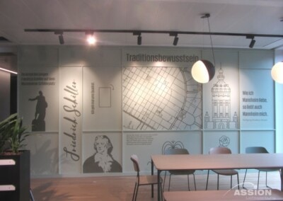 Moderner Büroraum mit einer Wand mit deutschen Texten, einem Porträt von Friedrich Schiller, Architekturzeichnungen und stilisierten Designelementen. Tische und Stühle stehen im Vordergrund.