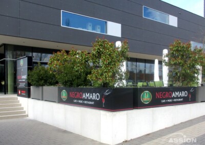 Sitzbereich im Freien des Negroamaro Café Music Restaurant, umrahmt von Pflanzen und Schildern, vor einem modernen schwarzen Gebäude.
