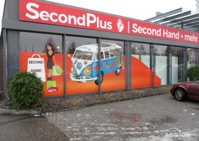 Eine Schaufensterfront mit der Aufschrift "SecondPlus Second Hand + mehr", großen Fenstern und einem Wandgemälde, das eine Frau beim Einkaufen und einen bunten Lieferwagen zeigt. Davor ist ein kastanienbraunes Auto geparkt.