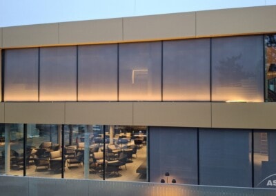 Modernes Gebäude mit großen Glasfenstern; im unteren Stockwerk befindet sich ein möblierter Essbereich mit Tischen und Stühlen, die Fenster im oberen Stockwerk sind undurchsichtig, aber eine sanfte Innenbeleuchtung ist sichtbar.