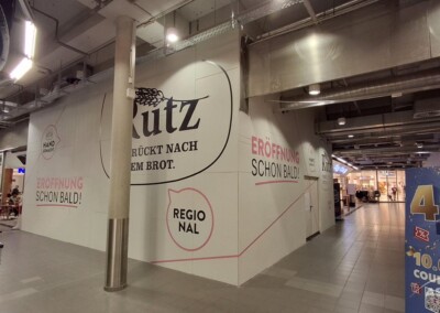 Ein Teil eines Einkaufszentrums, das gerade renoviert wird, mit Schildern, die die bevorstehende Eröffnung einer Kutz-Bäckerei ankündigen. Andere Geschäfte und Sitzgelegenheiten sind in der Nähe zu sehen.