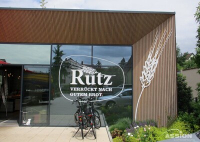 Eine moderne Bäckereifiliale mit großen Glasfenstern trägt das Logo "Rutz Verrückt nach gutem Brot" und eine Weizengrafik. Draußen sind zwei Fahrräder geparkt.